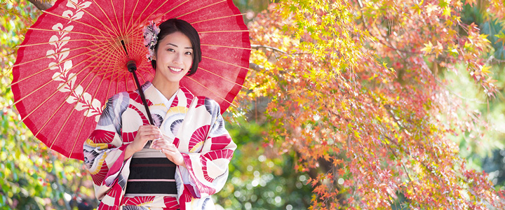 Eine junge Frau trägt einen kunstvoll gemusterten Kimono und einen roten Sonnenschirm. Umgeben von herbstlich gefärbten Ahornbäumen zeigt der Garten eine ruhige Atmosphäre.